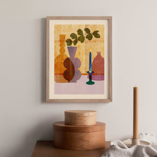 A framed still life print depicting colorful bottles and a candle holder, hanging on a wall above a wooden side table with a lamp and a cloth.